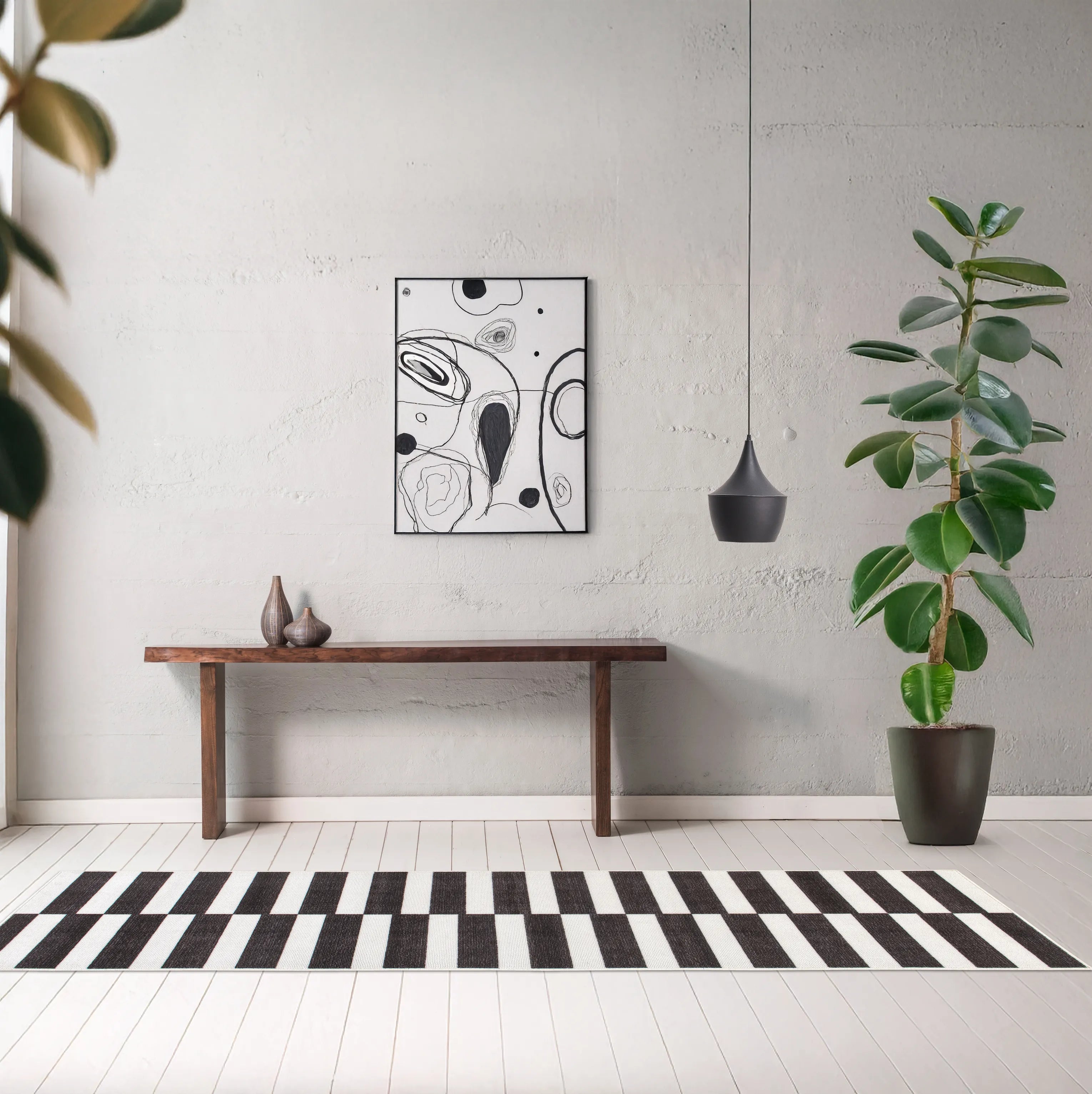 Tappeto Strider Black 80×250 cm a righe nere e panna alternate in corridoio con pavimento bianco, consolle in legno, quadro astratto bianco e nero, lampada a sospensione scura e pianta verde in vaso.