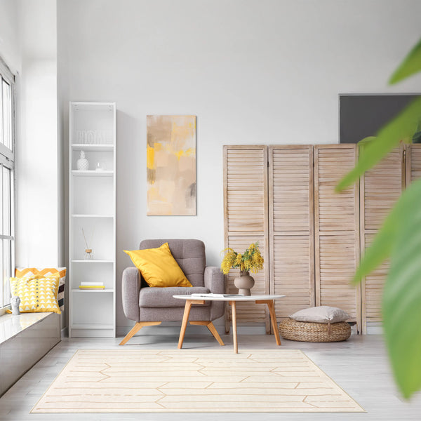 Tappeto Maira 130×190 cm con linee tribali beige su fondo crema, in soggiorno luminoso con poltrona grigia, cuscino giallo, separé in legno e piante; lavabile e asciugabile.