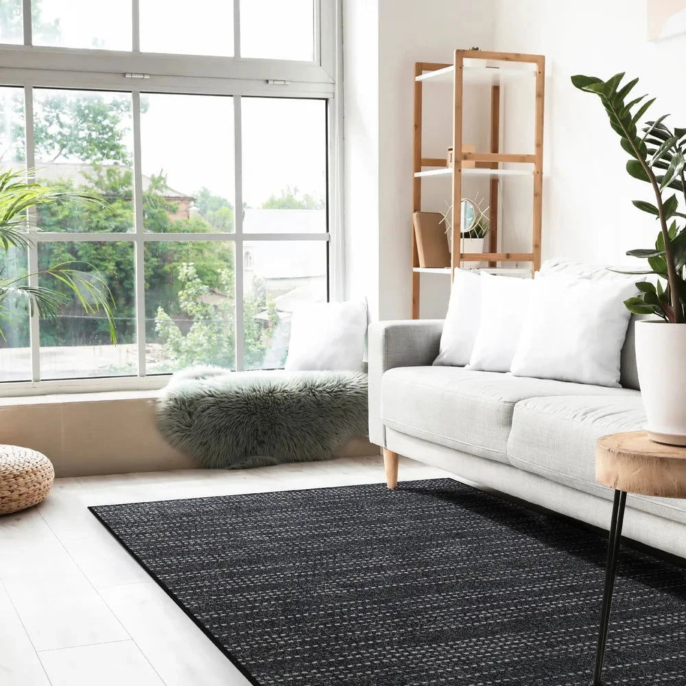 Tappeto Mono 170 × 240 cm Lindorug posato davanti a un divano chiaro e una panca con pelliccia sintetica, in un soggiorno luminoso con ampie vetrate.