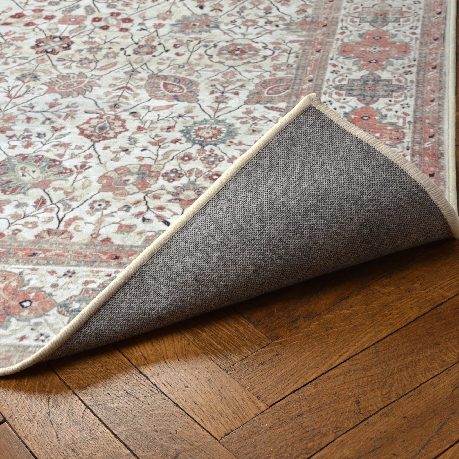 Vista del tappeto Sindhi con l’angolo sollevato che mostra il motivo floreale e la base antiscivolo e impermeabile; colori ecru e corallo, lavabile a 40°.